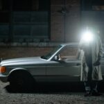 man in a trench standing beside the cat and holding a flashlight